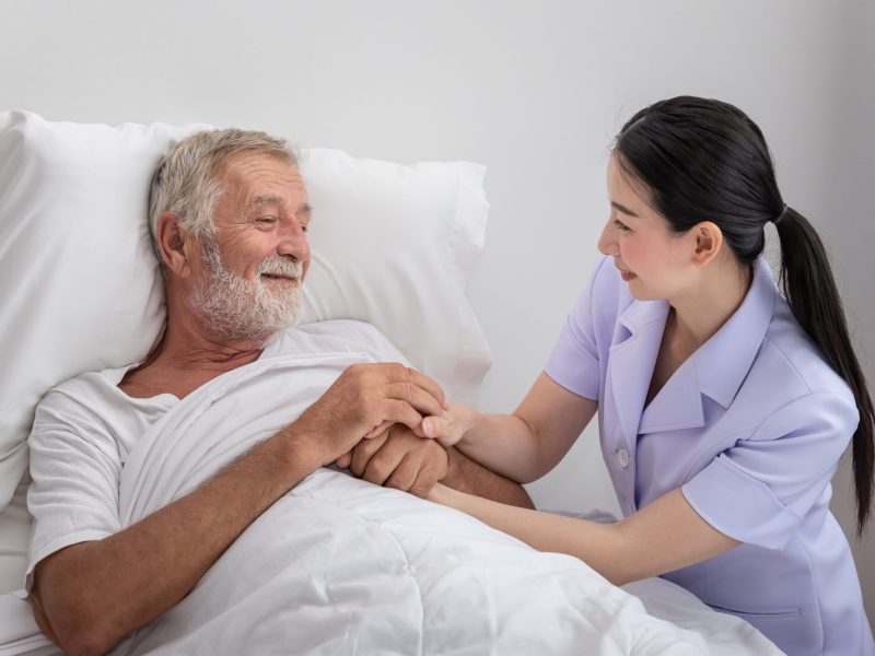 Happy nurse holding elderly man hands with blanket in bedroom at nursing home