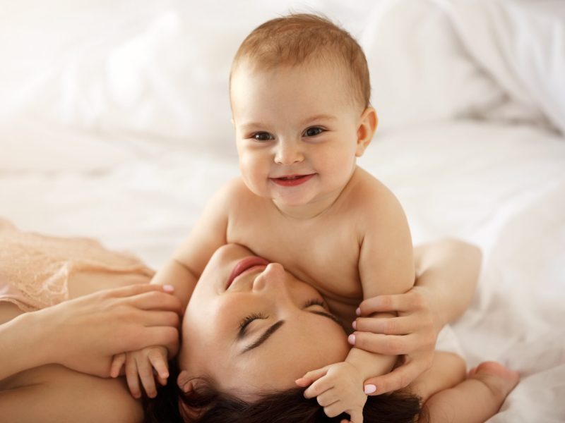 Young happy mother and her baby daughter smiling embracing lying in bed at home. Copy space.